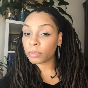 A Black woman with a V-shaped earring looks at the camera. She has long dreadlocks. Behind her, a bookcase, on which two potted plants sit.