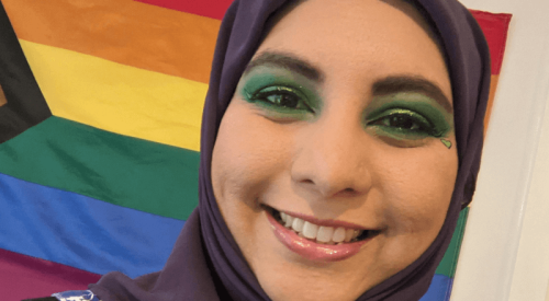 Woman of colour wears a hijab, a dress patterned with flowers and a badge that says Attraction beyond gender, in front of an inclusive Pride flag