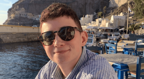 A white non-binary person wears sunglasses and a blue striped shirt and smiles at the camera. They are by a harbour with a mountain behind them.