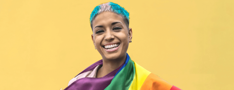 Person of colour with hair dyed in trans flag colours, draped in the rainbow flag, smiles at the camera