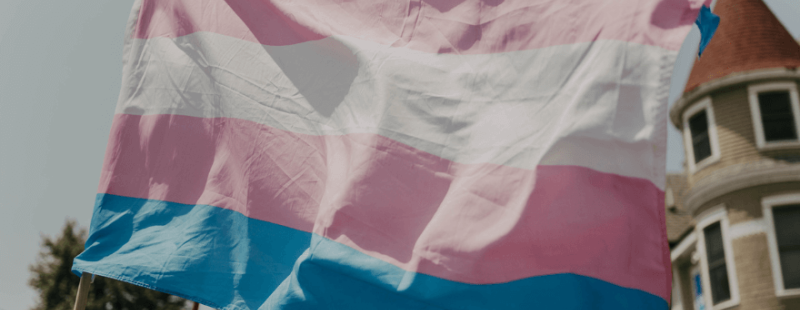 Trans flag in front of a rainbow flag in a sunny street