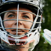 A person in lacrosse gear stands in a field, wearing a helmet and holding a lacrosse stick aloft.